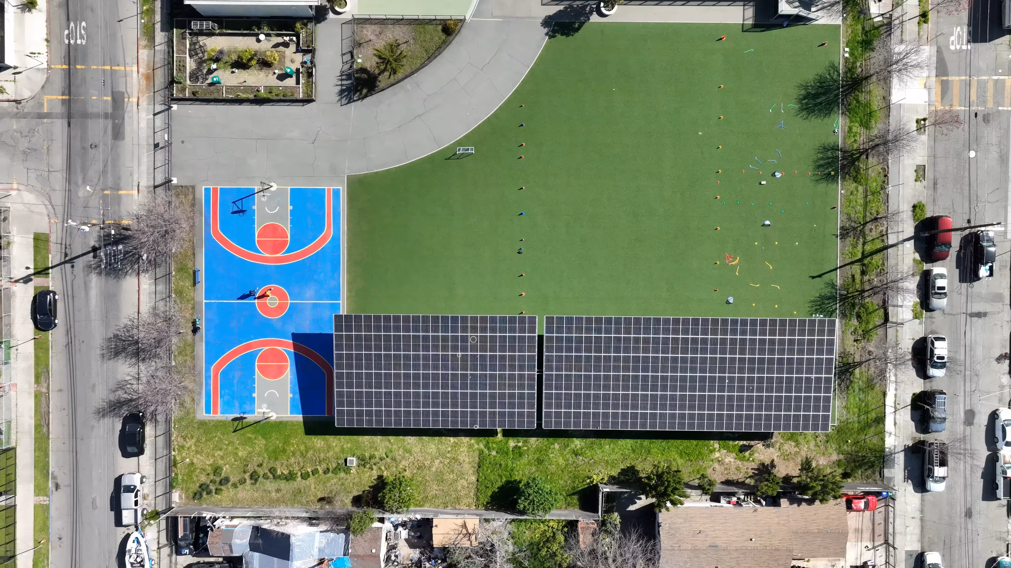 Aerial view of a blue and red basketball court next to a green field with cones and solar panels on a building roof.