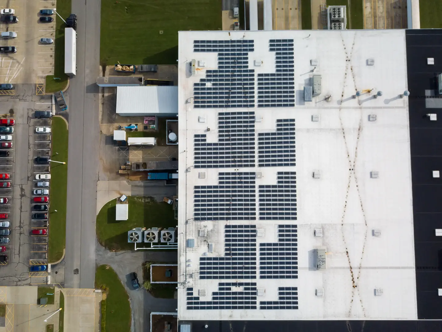 Aerial view of an industrial building roof with solar panels and adjacent parking lot with cars.