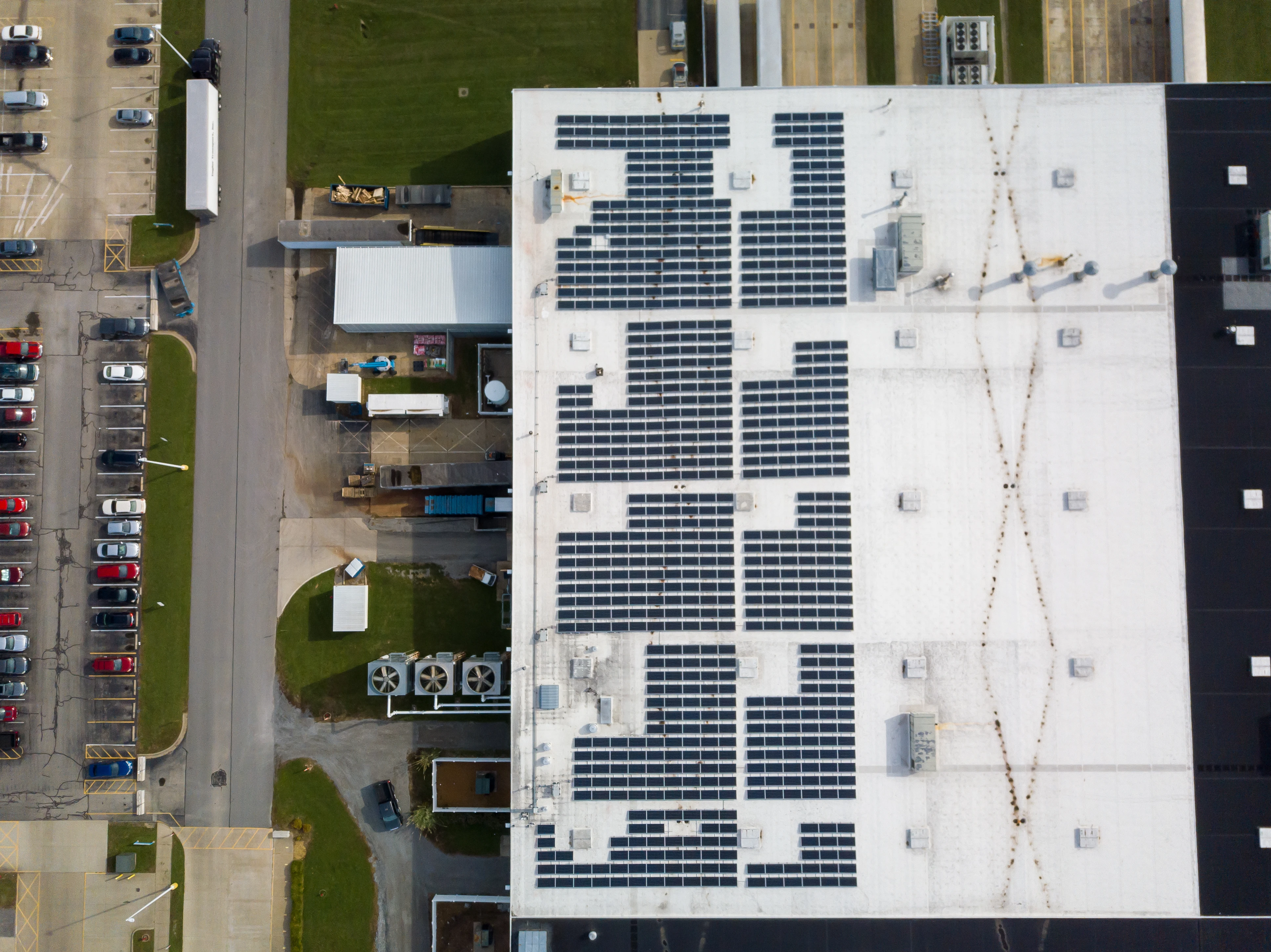 Aerial view of an industrial building roof with solar panels and adjacent parking lot with cars.