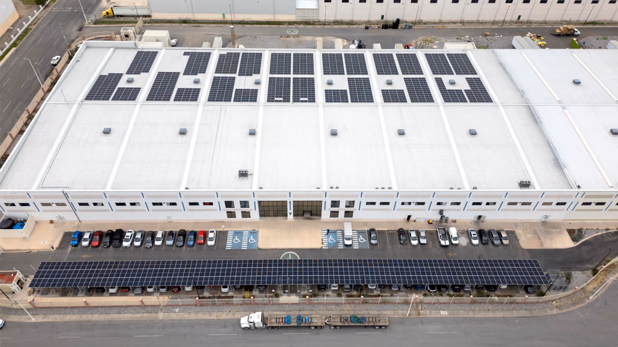 Aerial view of a large warehouse with solar panels on roof and a solar-covered parking area with parked cars.
