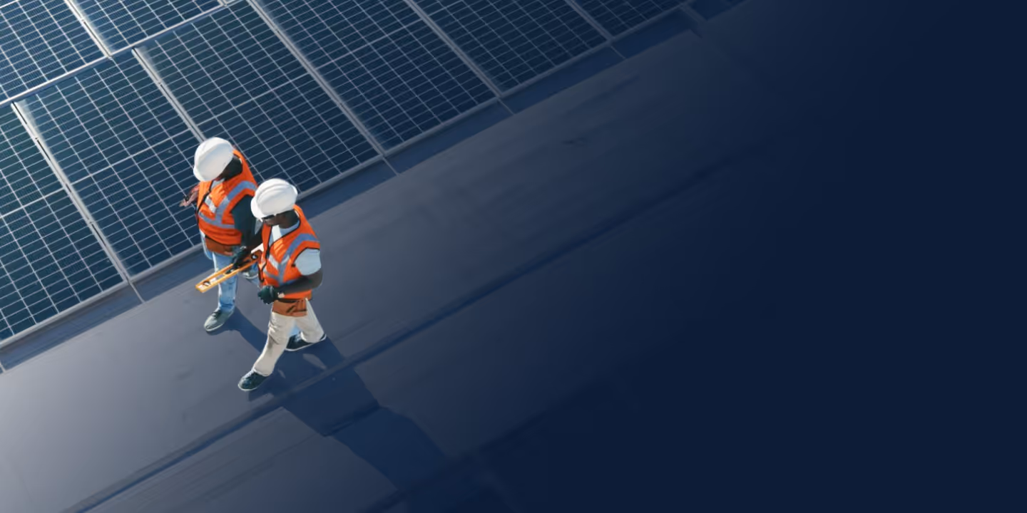 Two construction workers in hard hats and orange vests walking beside solar panels on a rooftop.