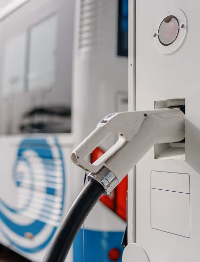 Close-up of a white electric vehicle charging plug connected to a charging station with a bus in the background.