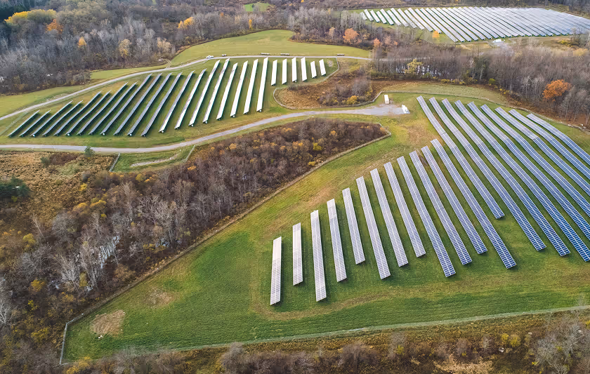 Aerial view of solar panels arranged in rows around grassy fields and wooded areas in autumn.