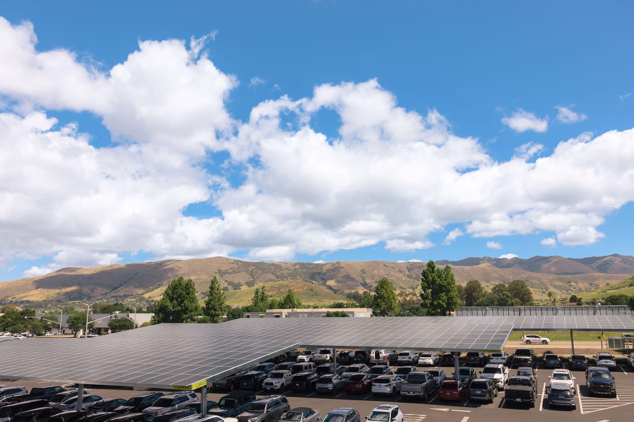 Solar panels covering a large parking lot full of cars with hills and partly cloudy sky in the background.