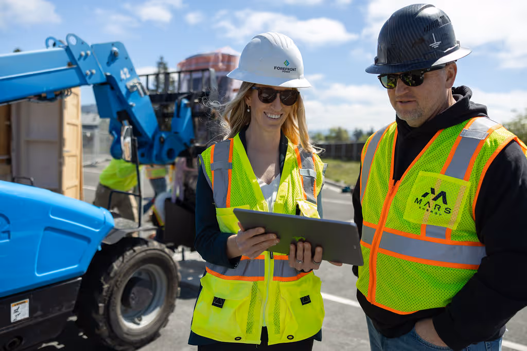 Two construction workers wearing safety vests and helmets reviewing a tablet on site near blue machinery.