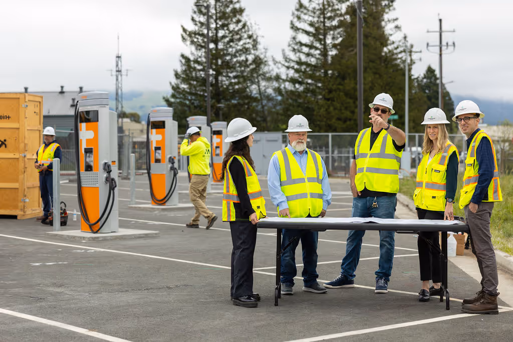 Group of construction workers in safety vests and helmets discussing plans outdoors near EV charging stations.