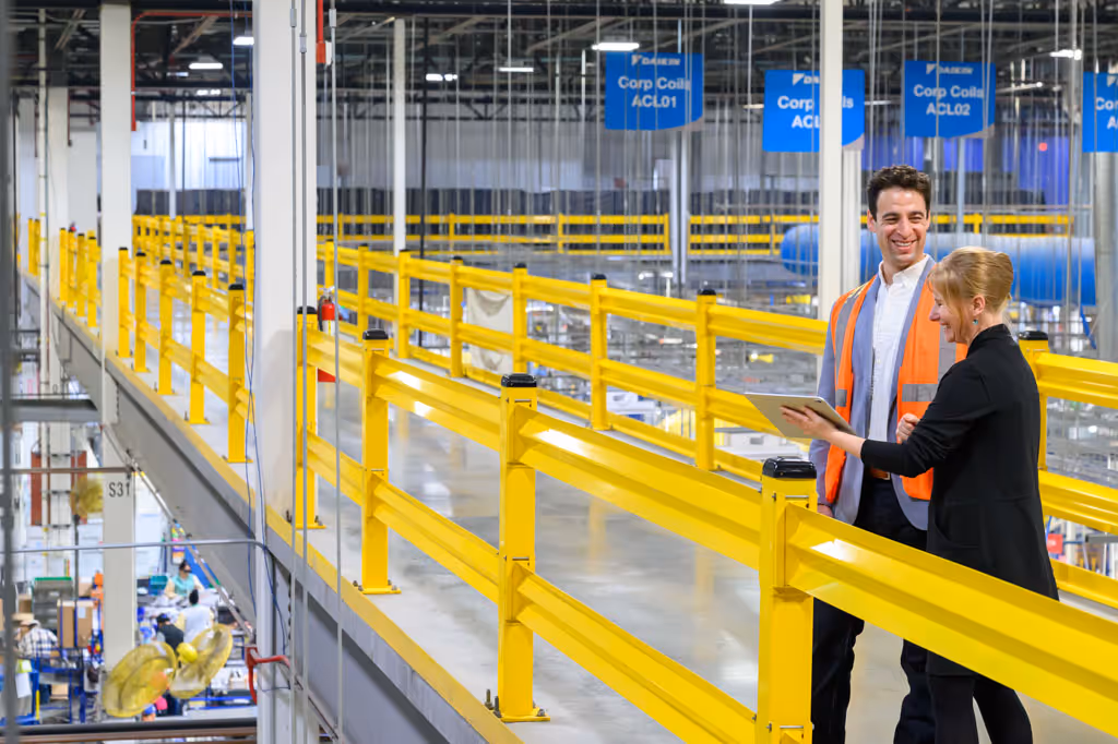 Two warehouse workers in safety vests review a tablet while standing behind yellow railings on a catwalk.