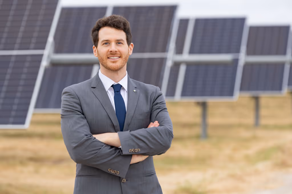 Man in gray suit standing with arms crossed in front of solar panels in a field.