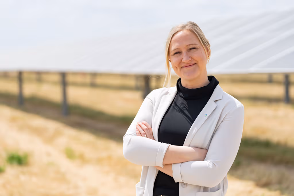 Smiling woman with blond hair standing with arms crossed in front of solar panels outdoors.