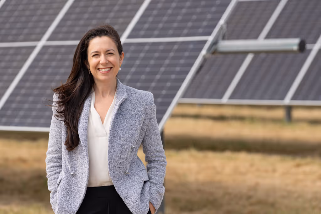 Smiling woman in white blouse and gray blazer stands in front of solar panels outdoors.