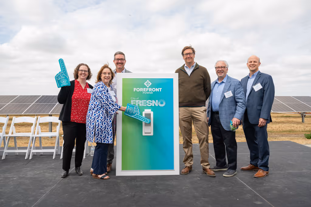 Six people stand onstage outdoors by solar panels with a large colorful light switch board.