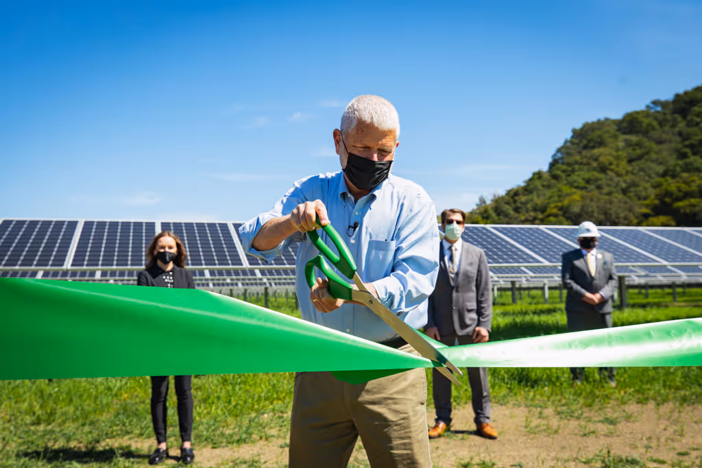 Man with gray hair and mask cutting green ribbon at solar panel installation ceremony outdoors.