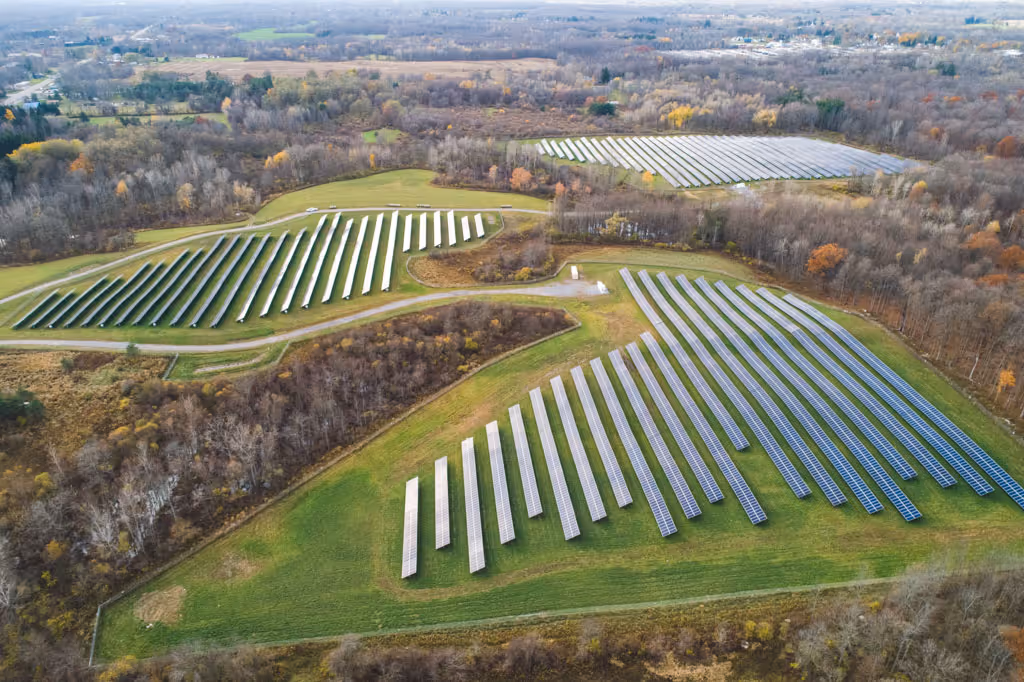 Aerial view of multiple fields with rows of solar panels surrounded by forest and farmland.