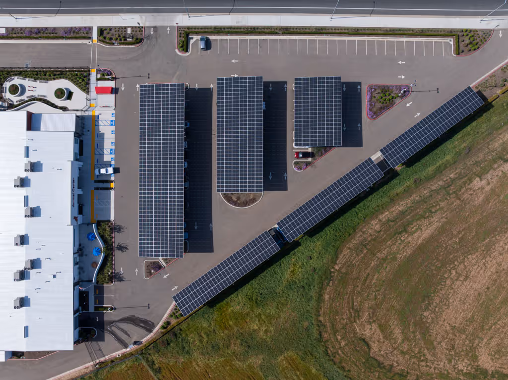 Aerial view of parking lot with solar panel canopies next to a large white roofed building and grassy area.