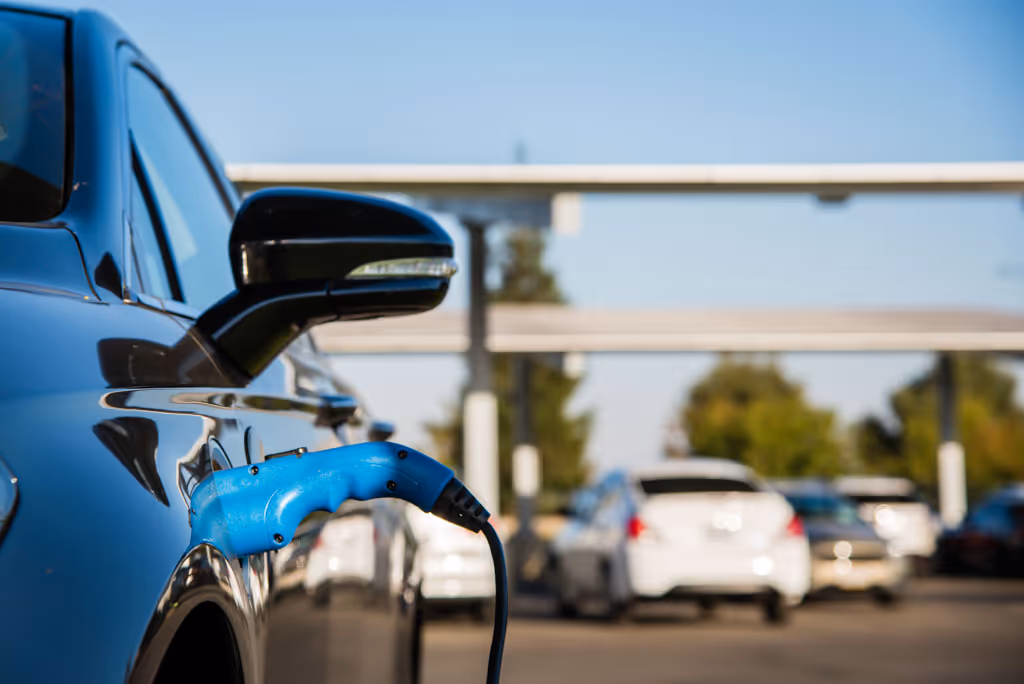 Black electric car charging with a blue plug at an outdoor parking lot under clear skies.