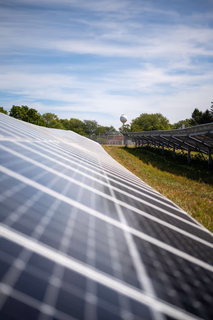 Rows of solar panels in a grassy field under a partly cloudy sky with trees and a water tower in the background.