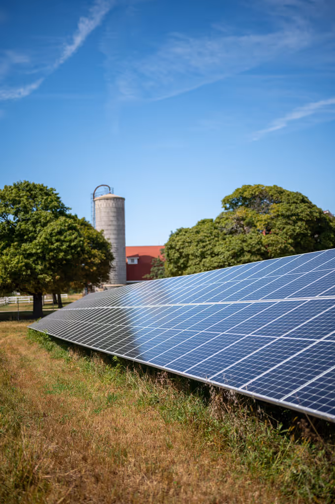 Solar panels installed on grass near trees and a farm silo under a clear blue sky.