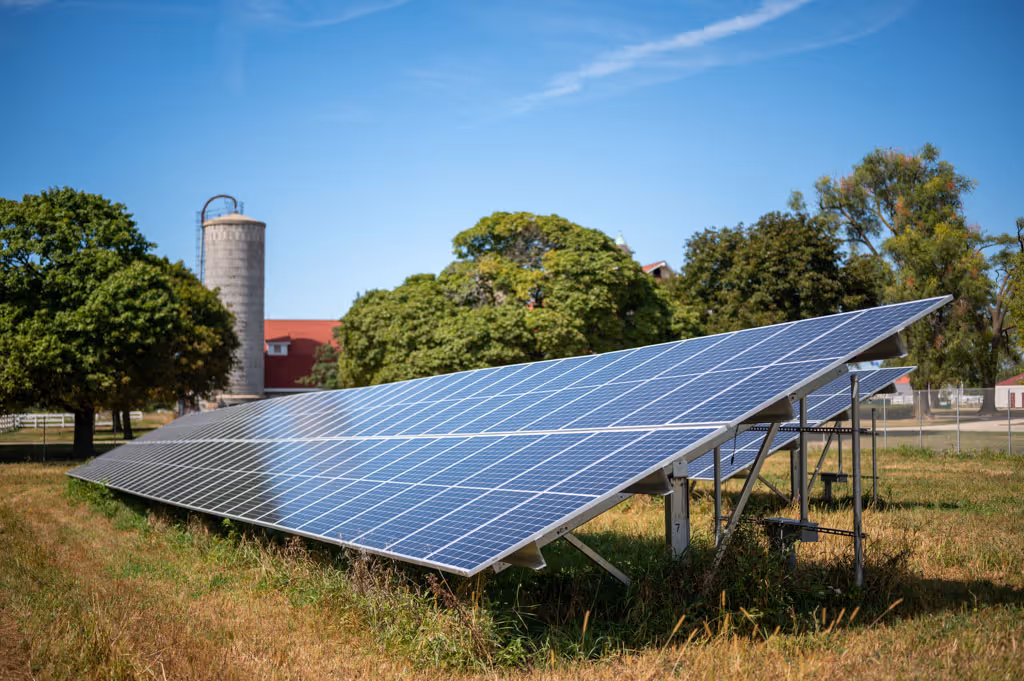 Solar panels installed on a grassy field with trees and a silo in the background under a clear blue sky.