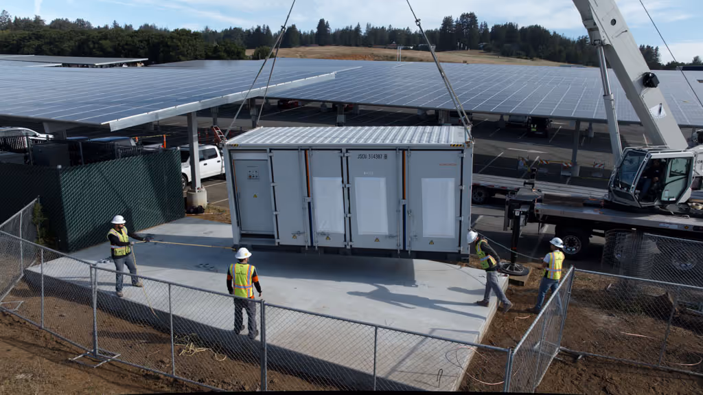 Workers in safety gear guide a large container being lifted near solar panel installations.