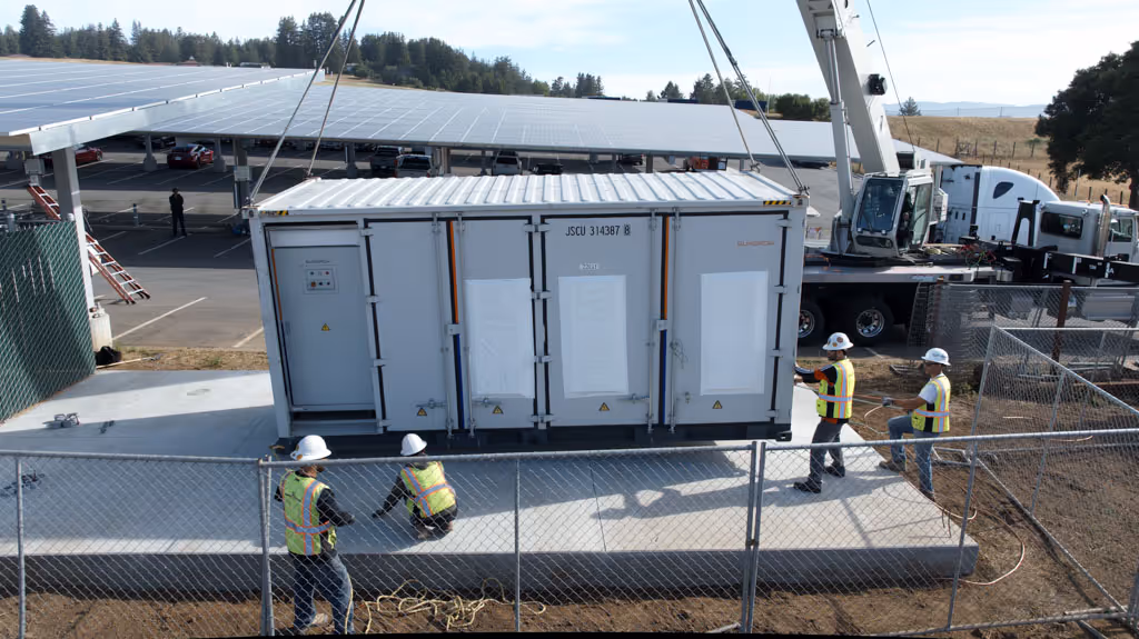 Workers wearing safety vests and helmets install a large electrical container near solar panels.