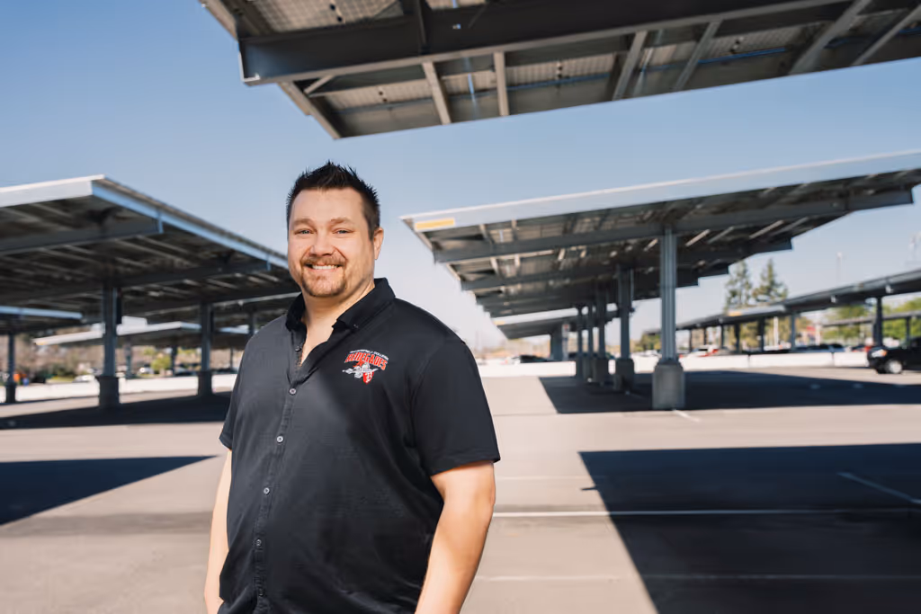 Smiling man in black shirt standing in a parking lot with solar panel canopies on a sunny day.
