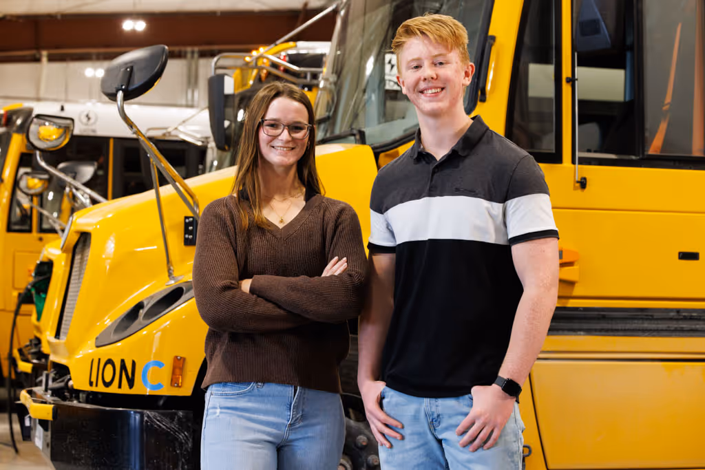 Smiling young woman and man standing in front of yellow Lion electric buses indoors.