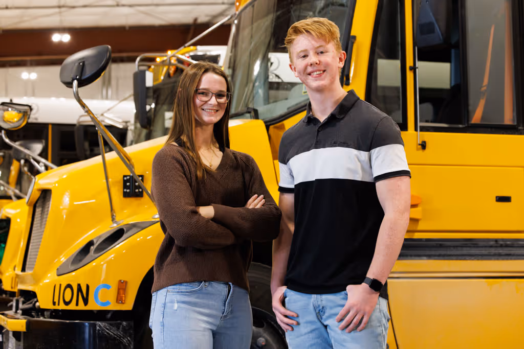 Two young people standing and smiling in front of a yellow Lion electric school bus.