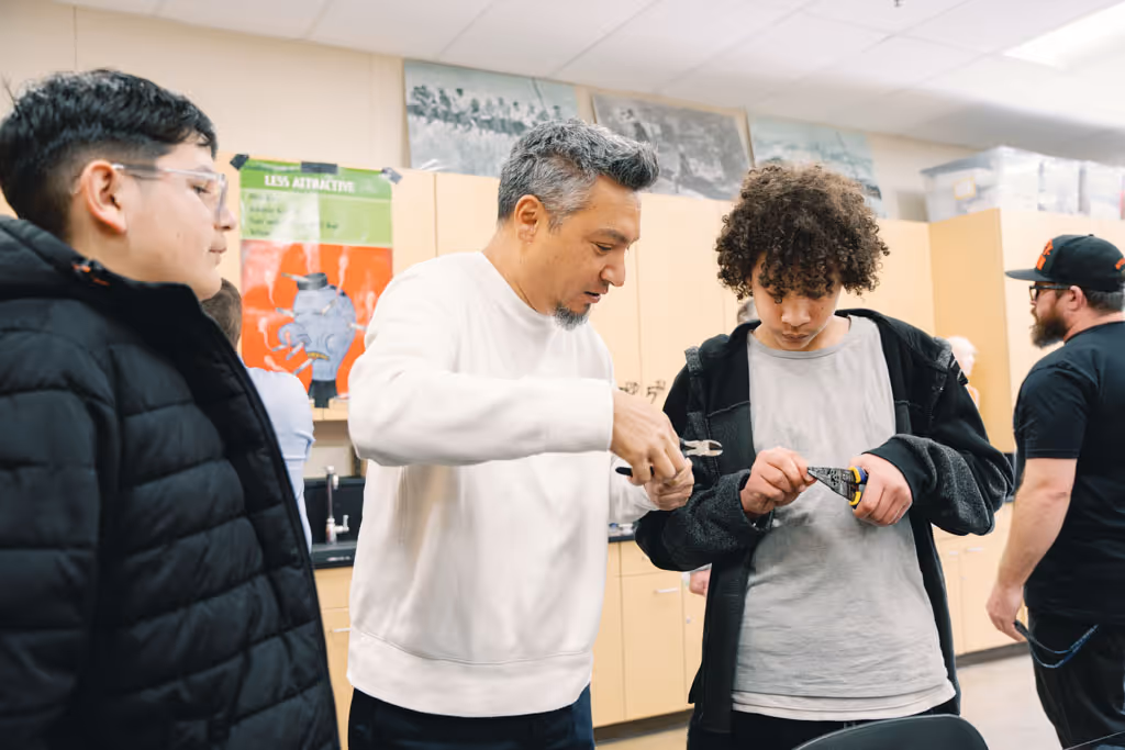 A man demonstrates tool use to teenagers in a classroom with cabinets and posters in the background.