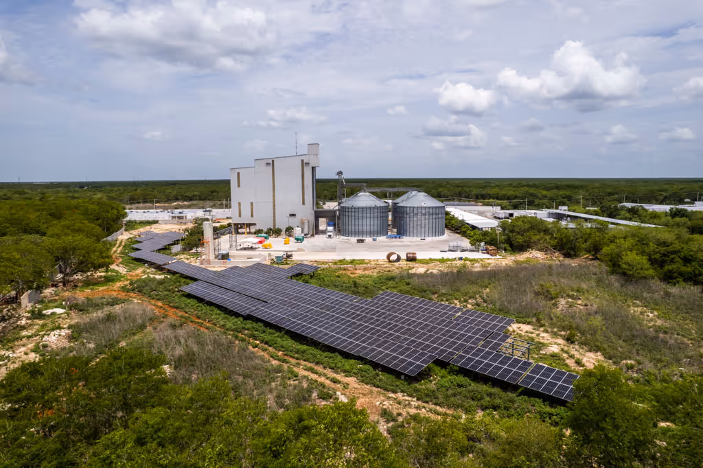 Aerial view of solar panels installed in front of an industrial building with large metal silos, surrounded by trees.