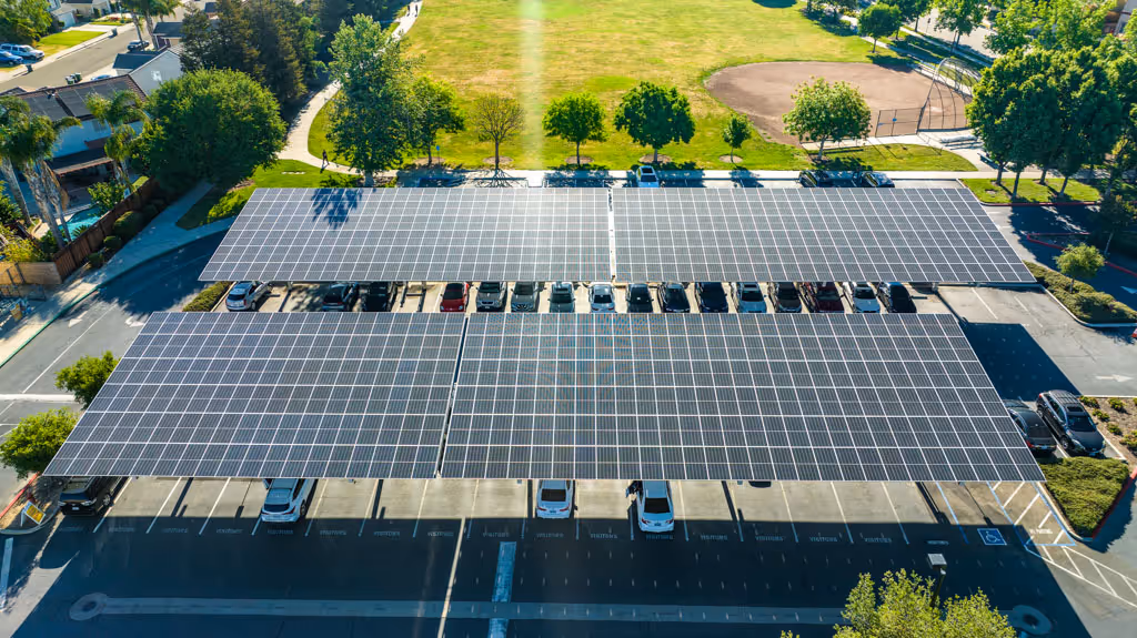 Aerial view of parking lot with cars under solar panel canopies beside a green park area.