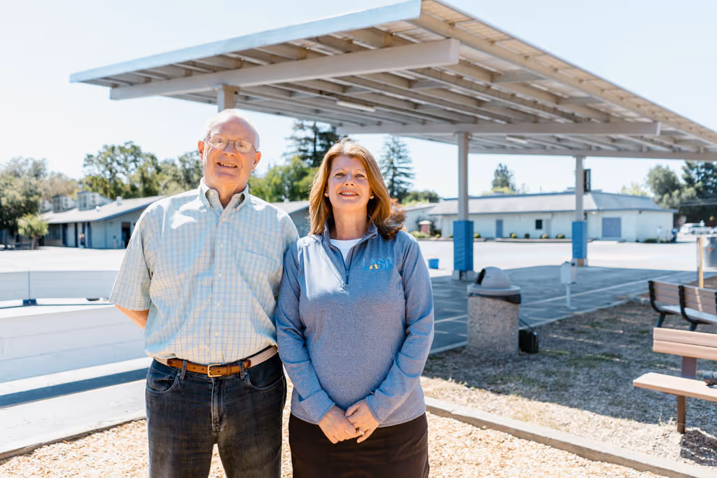 Smiling older man and woman standing outdoors near covered structure and buildings in daylight.