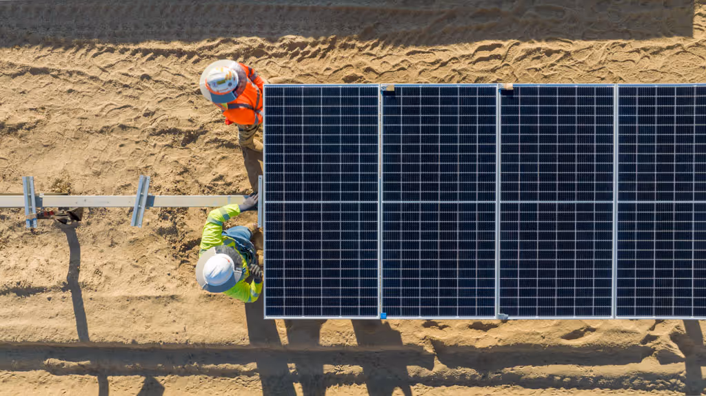 Two construction workers installing solar panels on a sandy ground viewed from above.