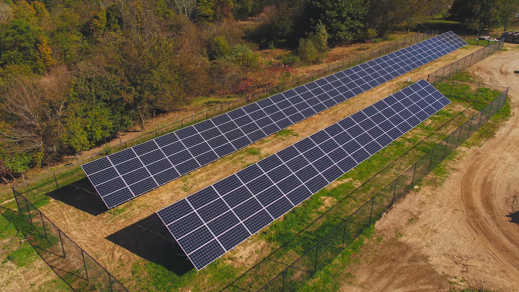 Two long rows of solar panels installed on a fenced grassy area adjacent to trees and dirt road.