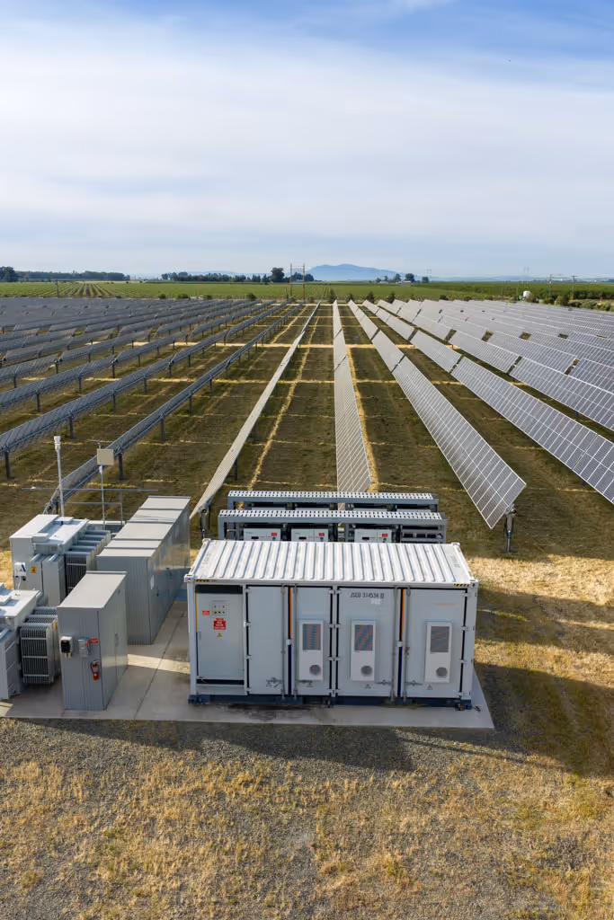 Rows of solar panels with electrical equipment and a container in the foreground under a partly cloudy sky.