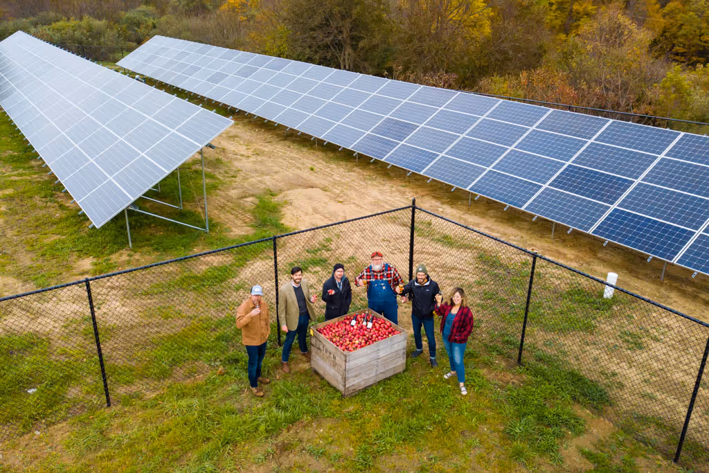 Six people standing around a wooden crate of apples near solar panels and a chain-link fence.