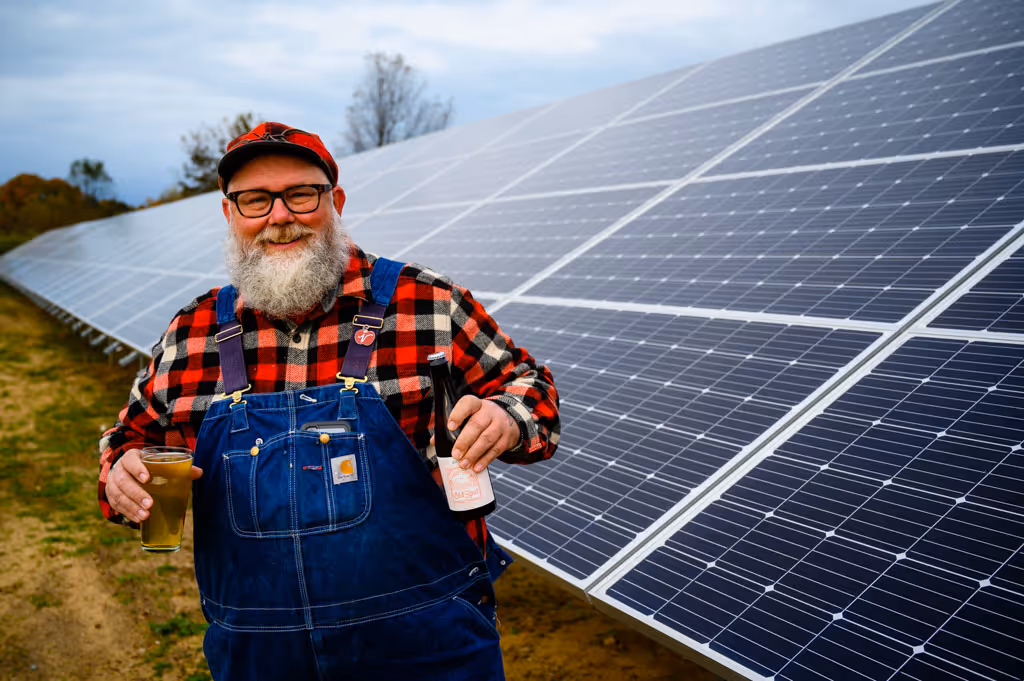 Smiling bearded man in red plaid shirt and overalls holding a beer glass and bottle by solar panels.