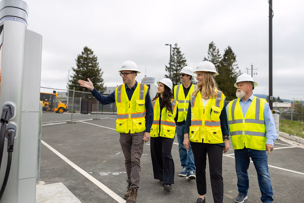 Five workers in yellow safety vests and white helmets inspecting outdoor equipment at a parking area.