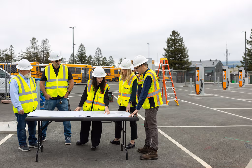 Group of construction workers wearing safety vests and helmets reviewing blueprints outdoors.