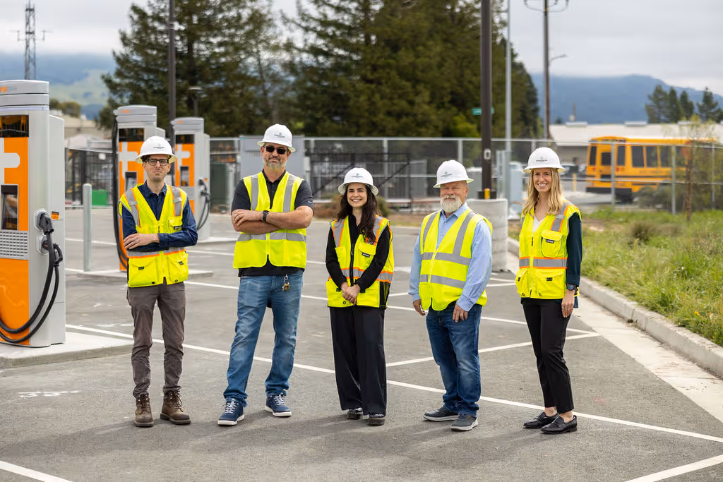 Five people wearing safety vests and helmets standing at an outdoor charging station area.