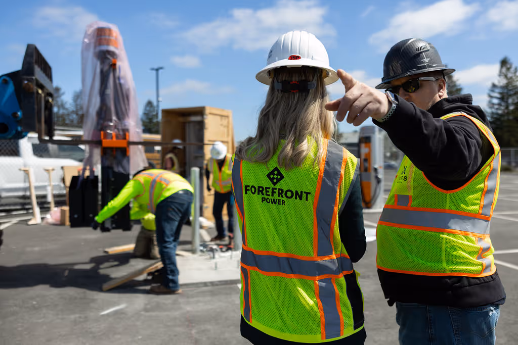 Two construction workers wearing safety vests and helmets, one pointing ahead, others working in background.