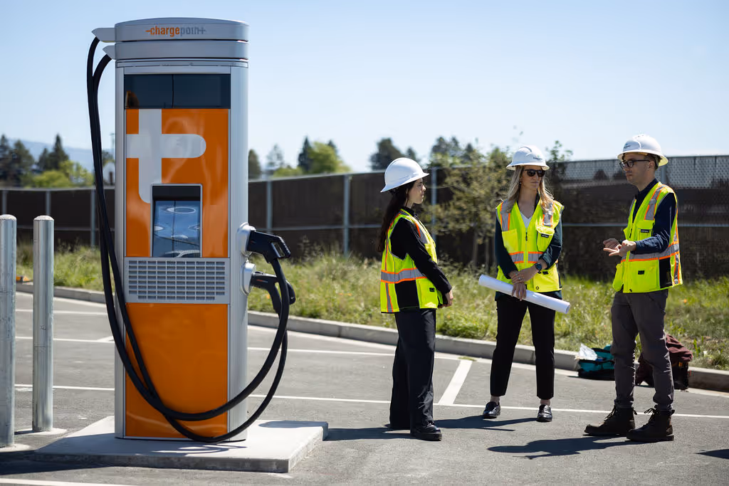Three construction workers with helmets and vests talking near an electric vehicle charging station.