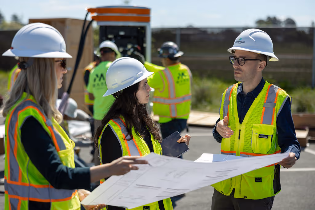 Three construction workers wearing hard hats and safety vests reviewing blueprints outdoors.