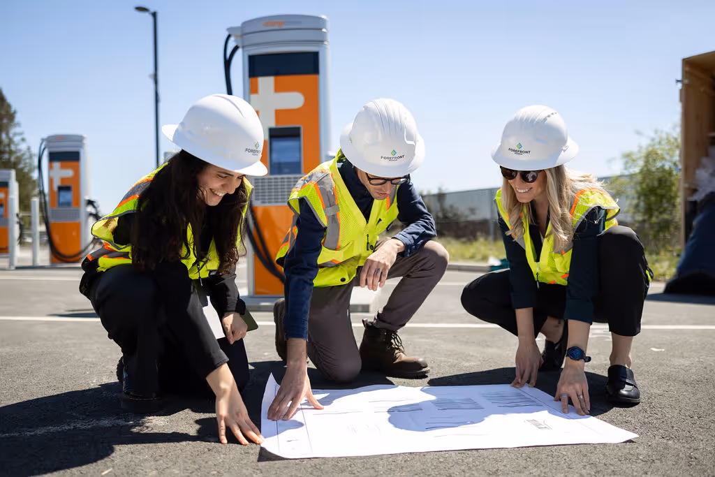 Three workers in safety vests and helmets reviewing plans at an electric vehicle charging station.