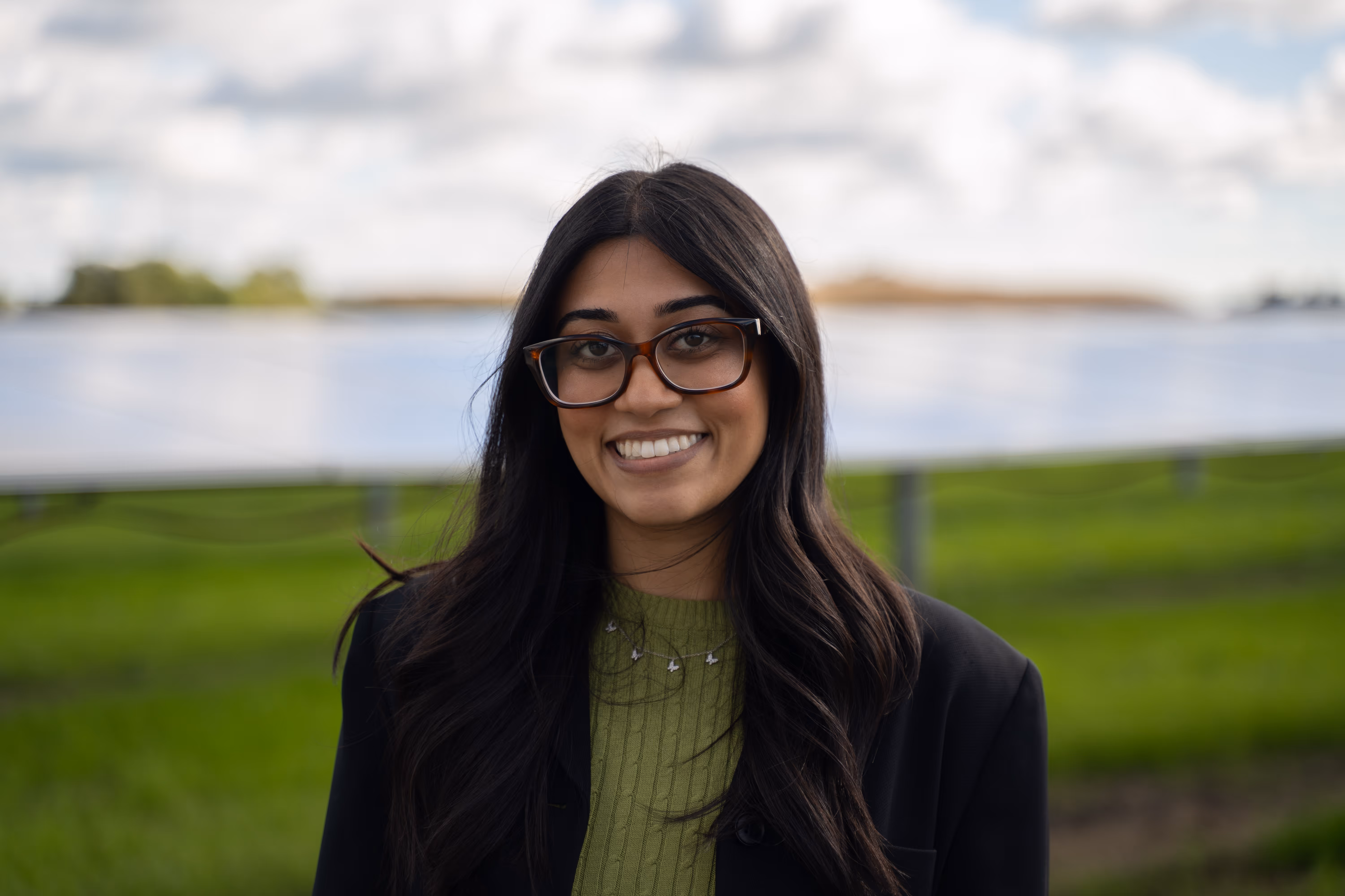 Smiling woman with long dark hair and glasses standing outdoors with green grass and solar panels behind her.