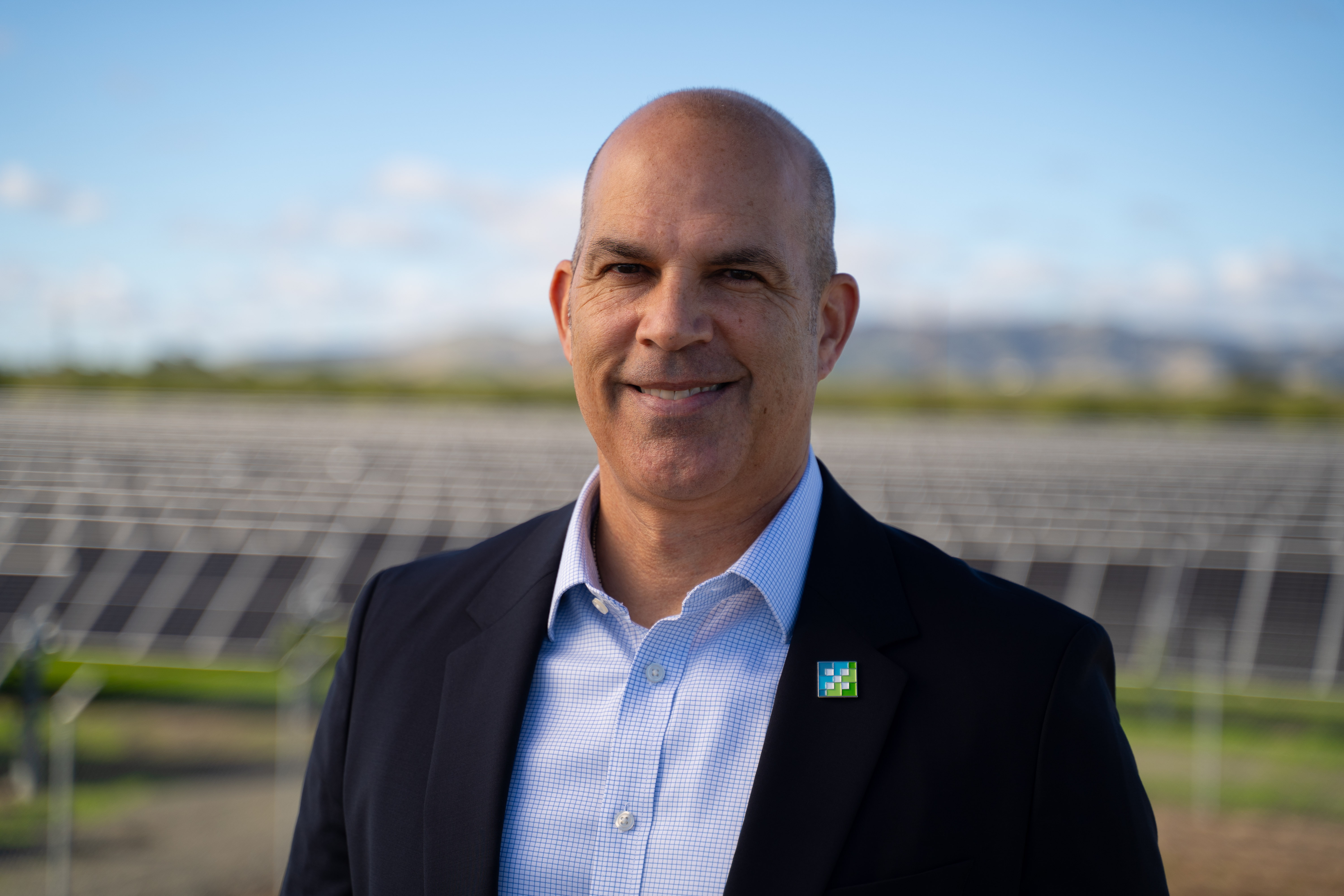 Smiling bald man in a blazer and checkered shirt standing in front of solar panels outdoors.