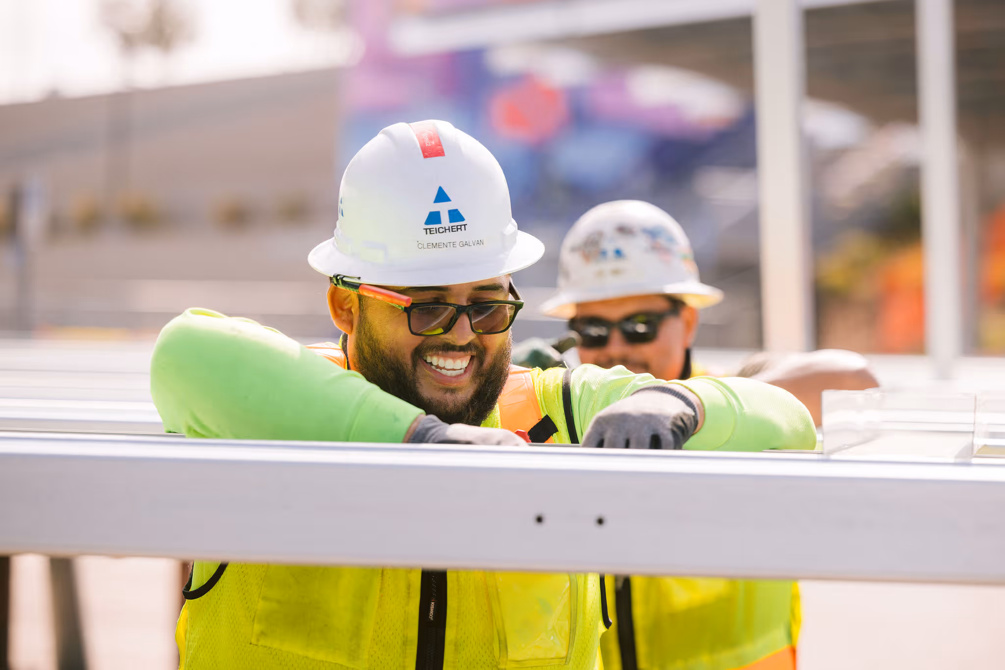 Two construction workers wearing hard hats and yellow vests assembling a metal structure outdoors.