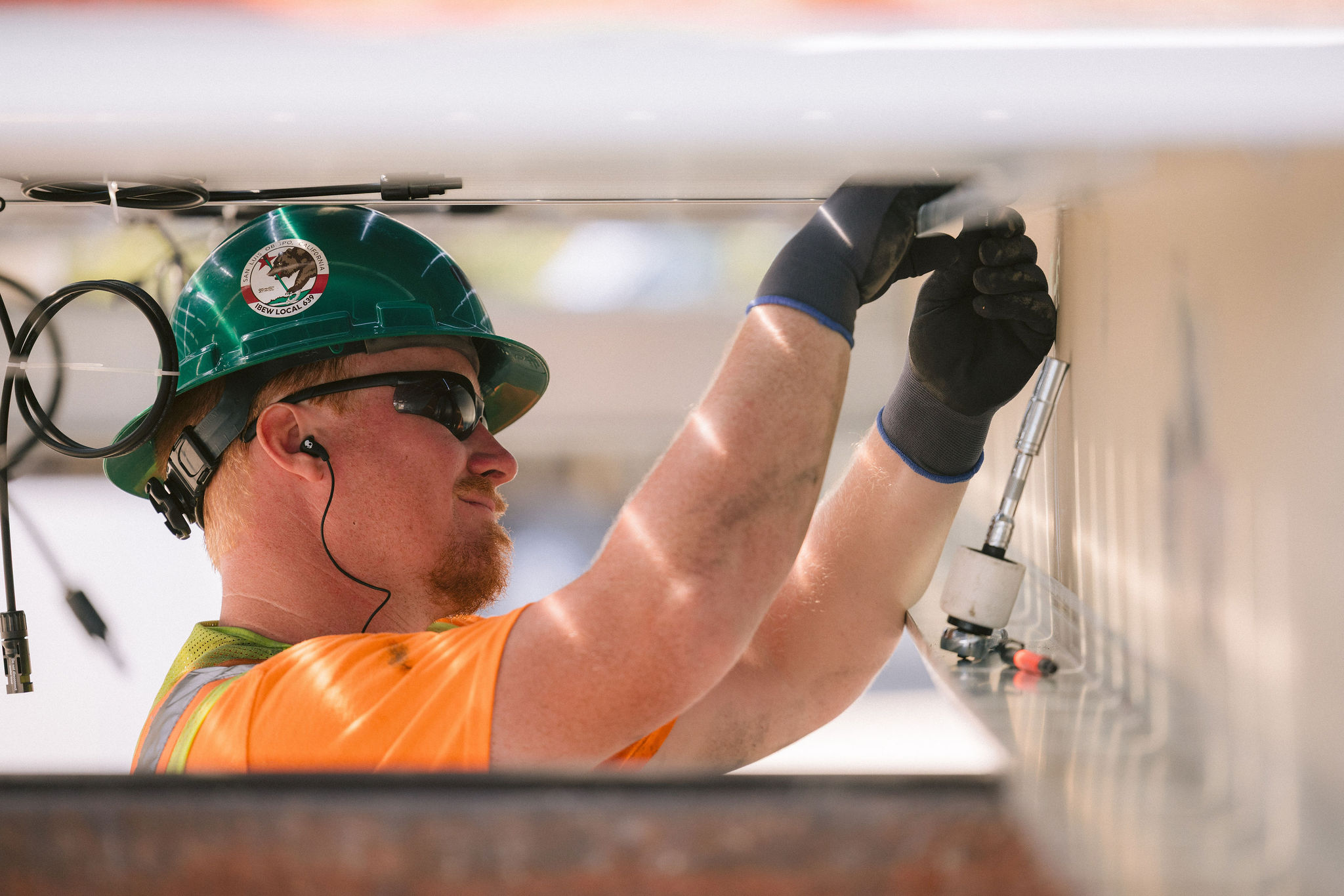Construction worker with green helmet and gloves installing equipment overhead.