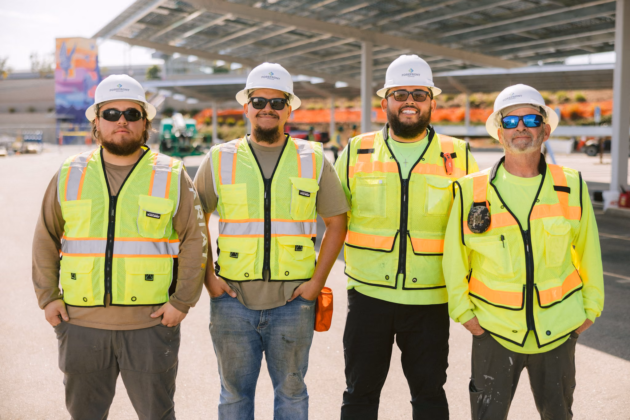 Four construction workers standing side by side wearing safety vests, hard hats, and sunglasses outdoors.