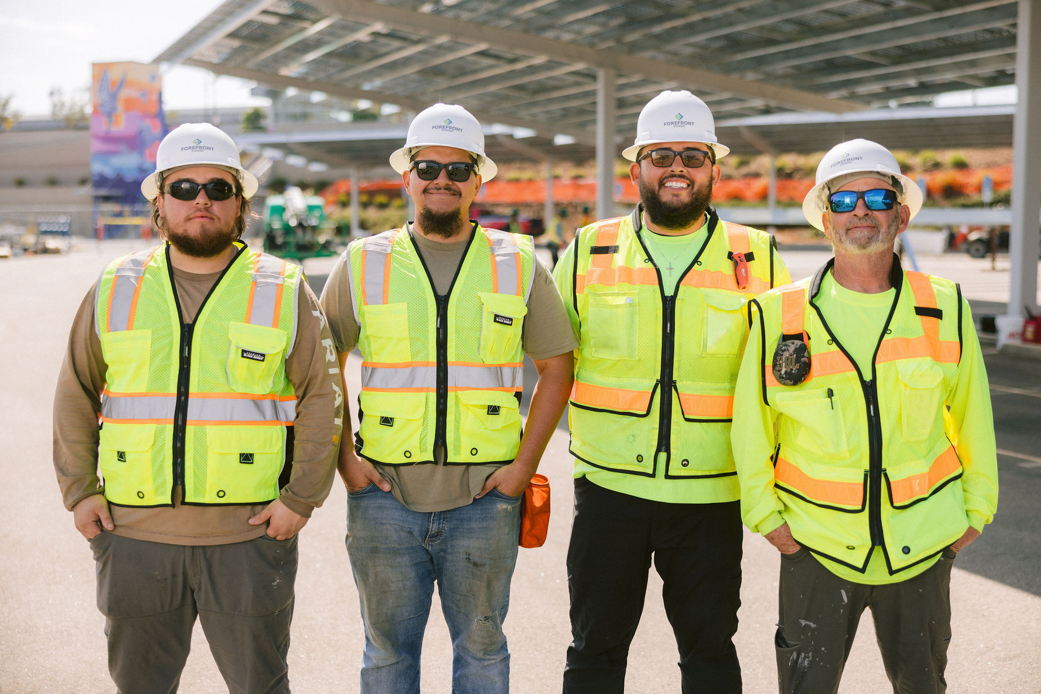 Four construction workers standing side by side wearing safety vests, hard hats, and sunglasses outdoors.