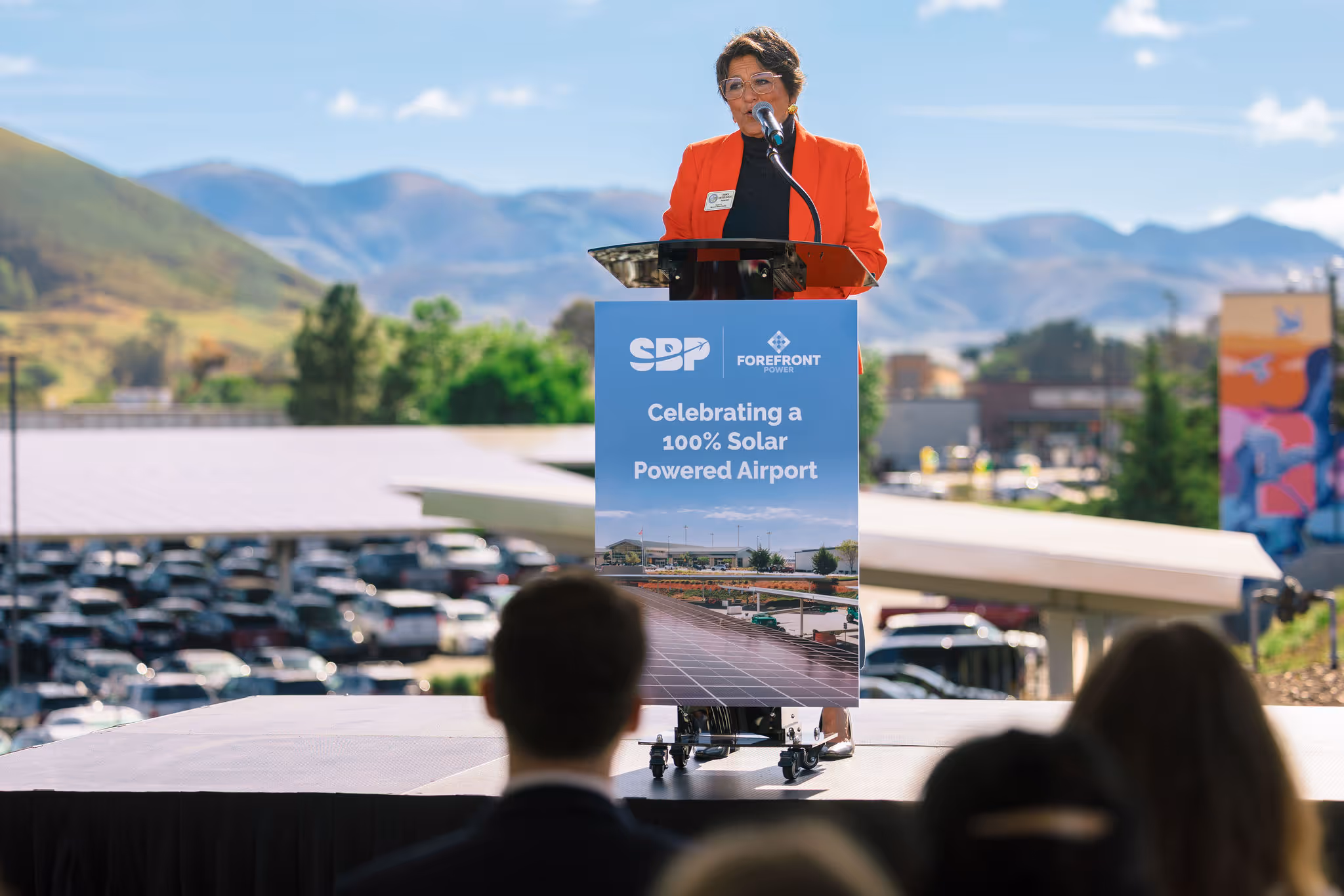 Person in orange jacket speaking at podium with sign about 100% solar powered airport celebration.